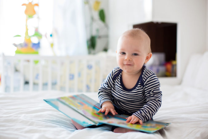 Lächelndes Baby sitzt mit Buch auf dem Schoß in seinem Kinderzimmer