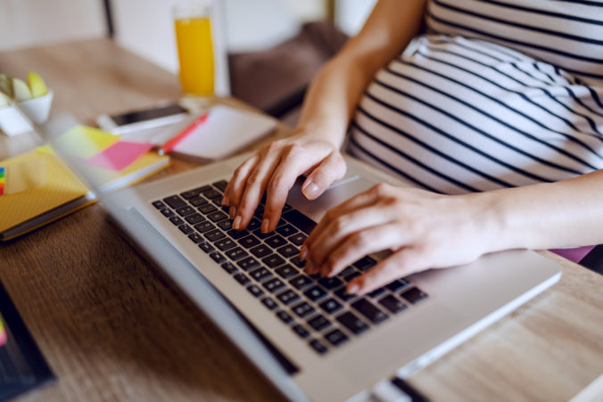 Schwangere Frau sitzt an einem Tisch und schreibt etwas auf dem Laptop