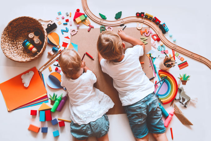 2 Kinder liegen auf dem Boden und malen. Um sie herum liegt eine Vielzahl von verschiedenen Spielzeugen